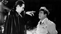Black and white scene with a person in a dark cape pointing at another person in a uniform.
