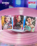 Three collectible character cards from KPop Demon Hunters stand on a pink vinyl record. The cards feature stylized illustrations of characters with the 'Hunter x' logo at the bottom.