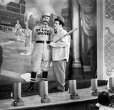 Two men on a stage with one in a St. Louis Wolves uniform and a baseball bat, with a cityscape backdrop.